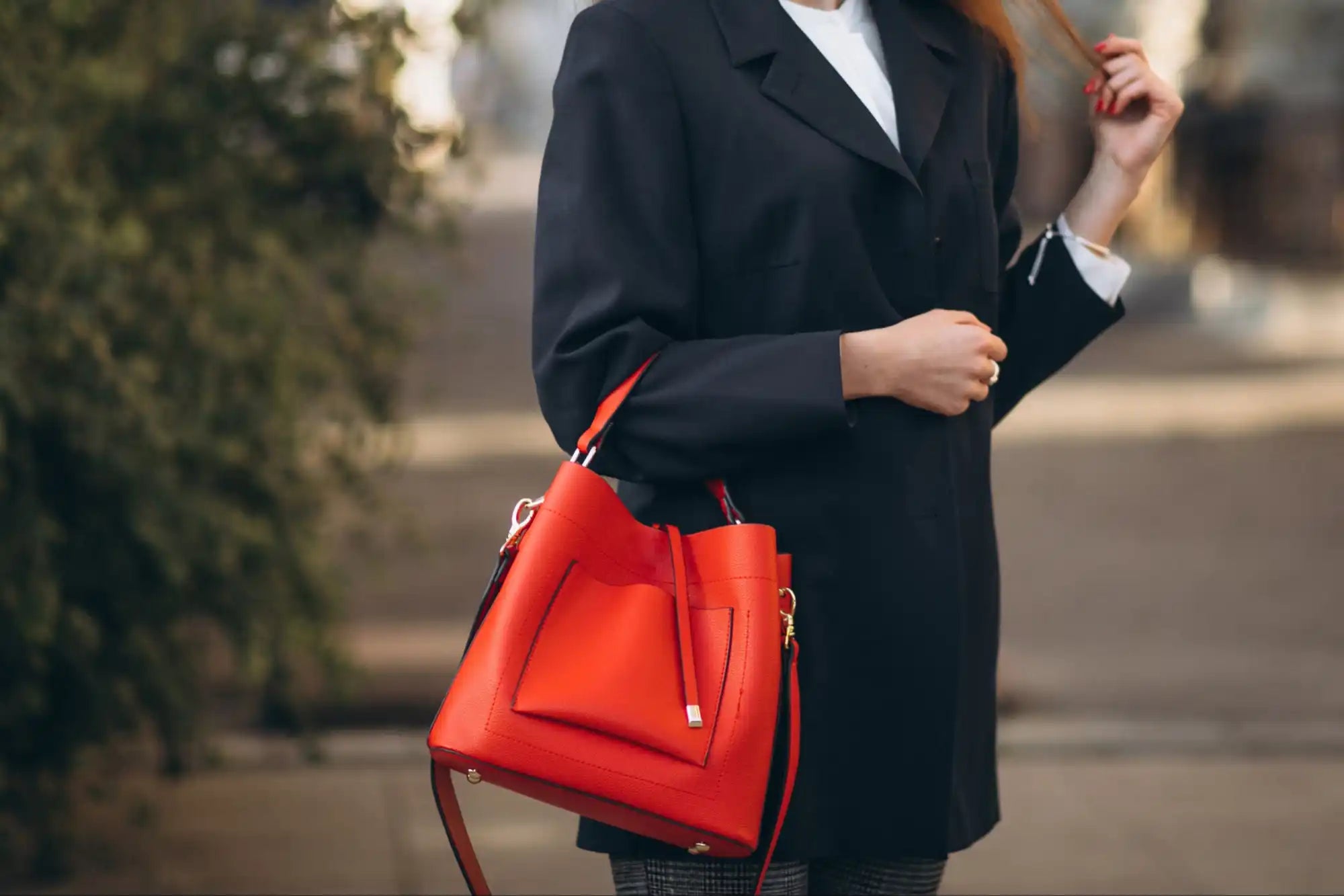 A person wearing a black blazer and white shirt carries a vibrant red leather bucket bag with an exterior pocket and drawstring closure.