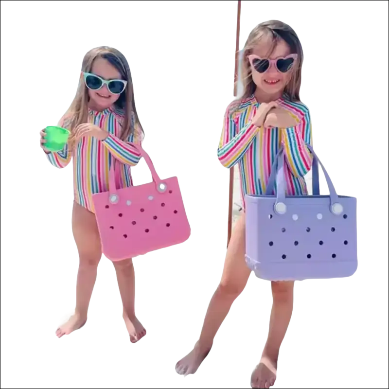 Two young girls in colorful striped swimsuits standing on a beach with sunglasses and tote bags.